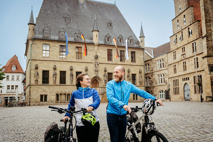 Friedensroute: Historisches Rathaus in Osnabrück. Friedensroute: Historisches Rathaus in Osnabrück mit zwei Radfahrenden, die ihre Fahrräder schieben, davor. .